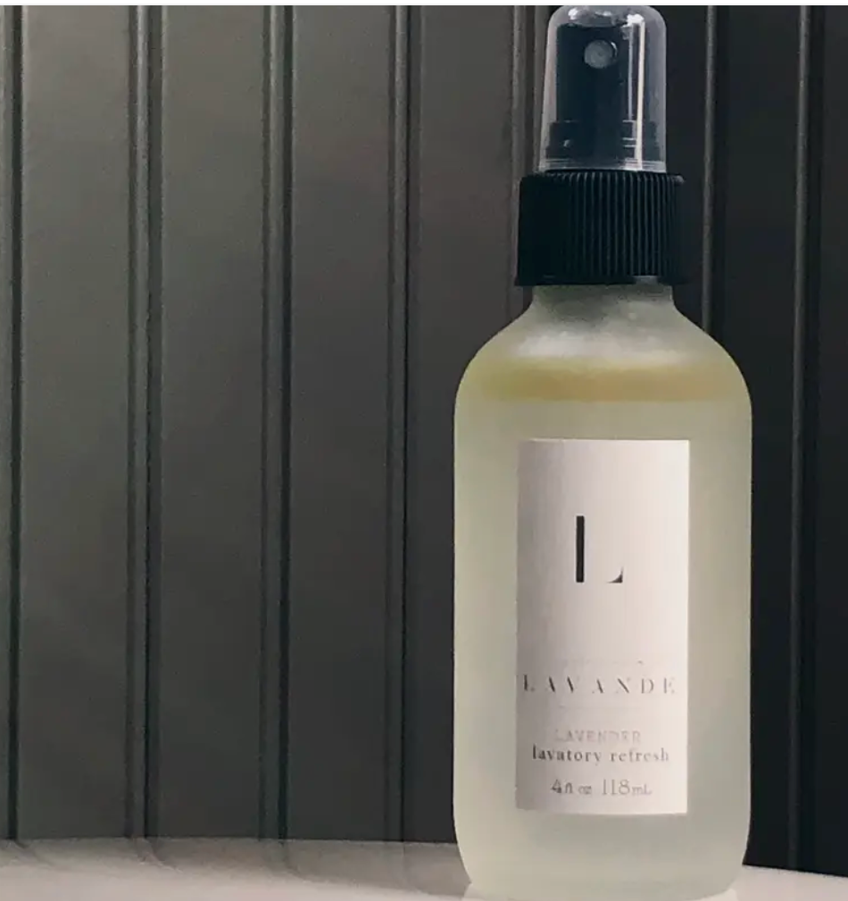 Pictured is a bottle of Lavande Room Spray sitting on a table with a black bead board wall in the background.