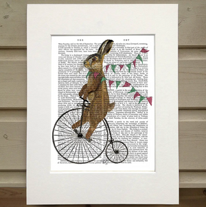 There is a page from a book framed with mat. Printed over the words on the page is a bunny riding an old fashioned bicycle that has a large wheel in the front and a small wheel in the back. Around the neck of the rabbit is a bunting with green and red flags, as if the bunny is crossing a finish line or wearing it like a scarf.