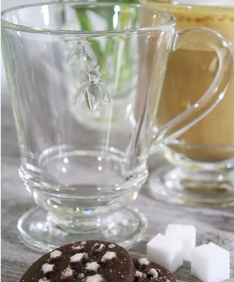 A bee coffee mug sits on a table next to 3 sugar cubes and 2 chocolate desserts. There is a Bee Coffee Mug by La Rochere sold at The Hare & The Hart.