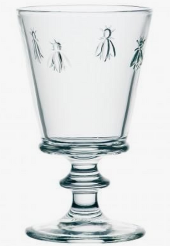 A clear water glass with four bees embossed on it. It has a decorative stem and is cylindrical in shape. This is a Bee Water Glass by La Rochere sold at The Hare & The Hart.