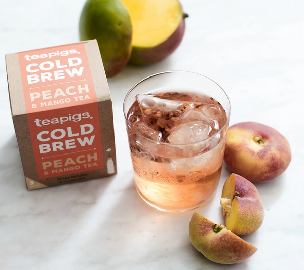 There is a package of Teapigs Cold Brew Peach and Mango tea. Next to it is a short glass of ice water with a teabag in it. Scattered around the package and the glass of tea are peaches and a mango.