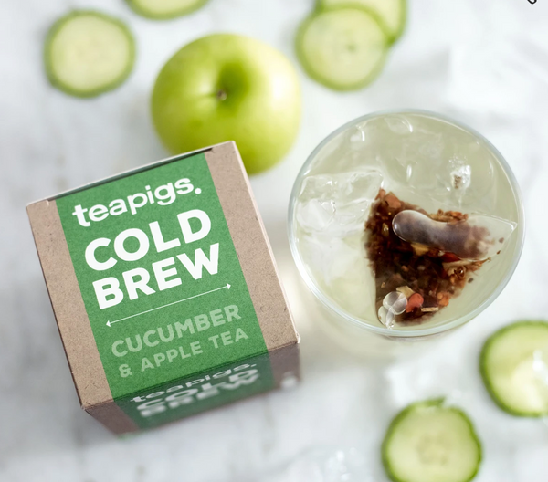From above, a glass of ice water with a teabag and a package of Teapigs Cold Brew Cucumber and Apple  is pictured. Sprinkled around them is an apple and slices of cucumber.