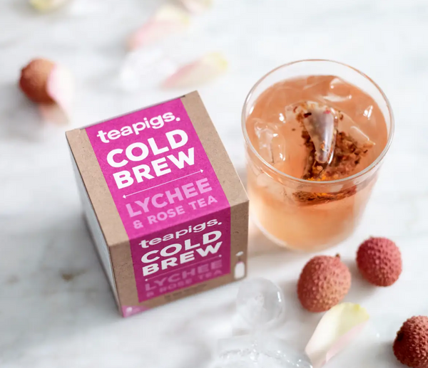 Pictured from above is a package of Teapigs Cold Brew Lychee and Rose tea next to a glass of ice water with a teabag in it. Surrounding the package and the glass are lychee fruits and rose petals.