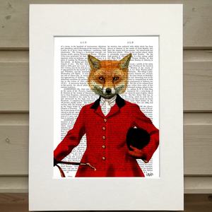 A page from a book is framed with mat. Printed over the page is a figure wearing an old hunting outfit with a red coat. In their left hand is the crop and tucked under their right arm is their riding helmet. Instead of the head of a person they have the head of a fox. This image is a portrait of the fox figure, cropped at the figure's waist. Pictured is a Fox Hunter Portrait Book Print by FabFunky Ltd sold at The Hare & The Hart.
