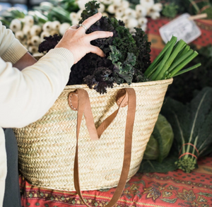 Pictured is a Double Handled French Market Basket by Medina Mercantile and sold at The Hare & The Hart. The basket is sitting on a table covered with a tablecloth. On the table are different vegetables, like at a farmer's market. A person is putting greens into the French market Basket.