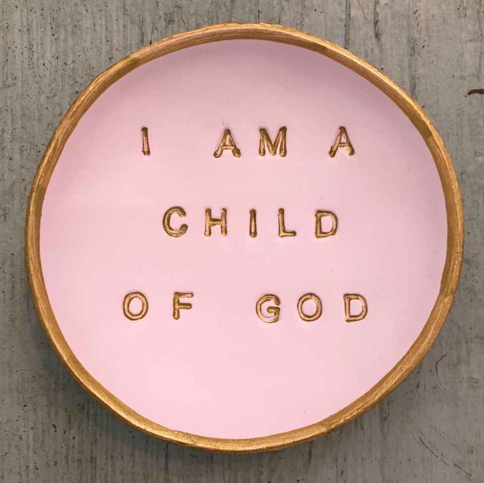 Pictued is a pink Blessing Bowl sold at The Hare & The Hart. It is a small, round, shallow bowl. Imprinted into the basin of the bowl are the words "I AM A CHILD OF GOD" in all caps, sans serif text. The text and the rim of the bowl are both decorated with gold leaf.
