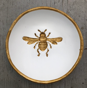 Pictured is a white Blessing Bowl sold at The Hare & The Hart. The bowl is small, round, shallow, and made of clay. Imprinted in the center of the basin of the bowl is a honey bee. The bee and the rim of the bowl are both decorated with gold leaf.