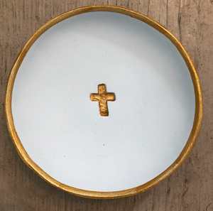 Pictured is a light blue Blessing Bowl sold at The Hare & The Hart. The bowl is small, round, shallow, and made of clay. It is painted light blue and has a small cross imprinted into the center. The cross and the rim of the bowl are both deocrated with gold leaf.