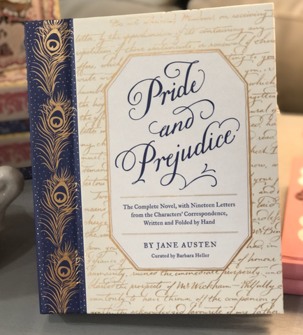Pictured is the book "Pride and Prejudice" by Jane Austen. The cover has gold embellishments of quotes from the book written in script as well as peacock feathers.