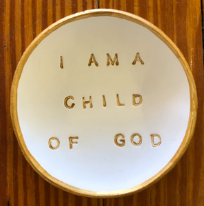 Pictured is a white blessing bowl with the words "I AM A CHILD OF GOD" in gold in the center of it. The rim of the clay bowl is also gold. Made in Oxford, MS by artist Carrie Cox and sold at The Hare & The Hart.