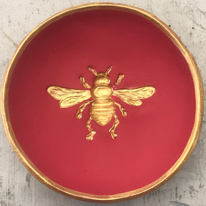 Pictured is a red blessing bowl with a gold bee in the center of it. The rim of the clay bowl is also gold. Made in Oxford, MS by artist Carrie Cox and sold at The Hare & The Hart.