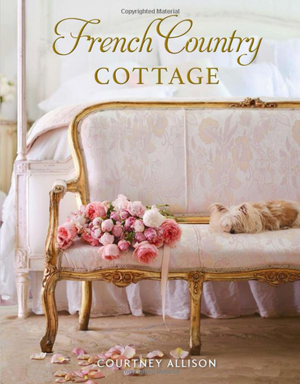 Pictured is the book "French Country Cottage" by Courtney Allison sold at The Hare & The Hart in Thomasville, GA. The cover photo is of a baroque style bench at the foot of a four poster bed. The bench is painted gold and has light pink, floral fabric upholstery. On the beach is a small dog sleeping and a bouquet of pink roses. Some of the petals have fallen onto the floor below. The bed in the background is all white. The picture has a light and airy feeling to it.