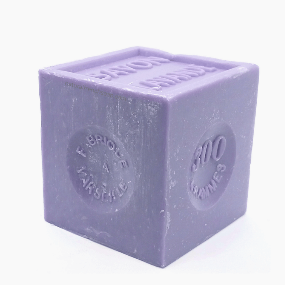 Pictured is a lavendar soap block by Savon de Marseille. The block is cut into a cube and is stamped with SAVON LAVANDE.