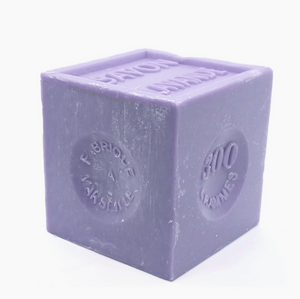 Pictured is a lavendar soap block by Savon de Marseille. The block is cut into a cube and is stamped with SAVON LAVANDE.