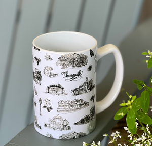 There is an average sized ceramic coffee mug. The mug has a white background and black Toile of Thomasville design.