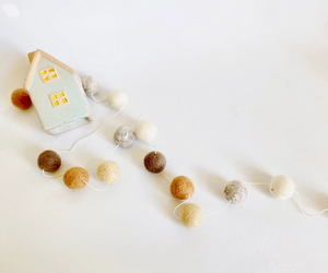 Pictured is a neutral wool felt ball garland. The garland lays on a white surface next to a ceramic sculpture of a cottage. The balls on the garland are various neutral colors, like tan, caramel, brown, white, and gray.