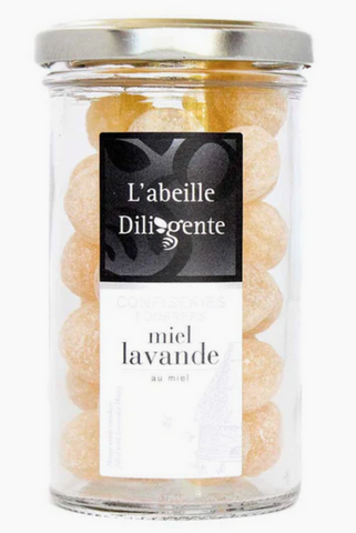 Pictured is a glass container of French Lavender Honey Filled Candy. The candies are yellow in color and have a sugary coating. The label reads "L'abeille Diligente miel lavande"