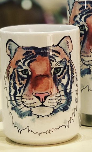 Pictured is a white coffee mug with a watercolor and pen & ink illustration of a tiger's face printed on it.